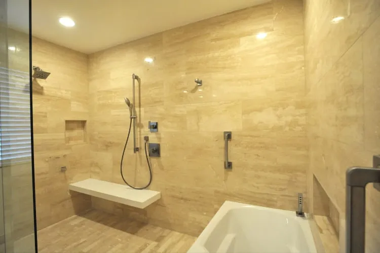 The finished walk-in shower: travertine walls, a floating stone bench, a fixed rain head with a separate hand shower, and grab bars beside the tub.