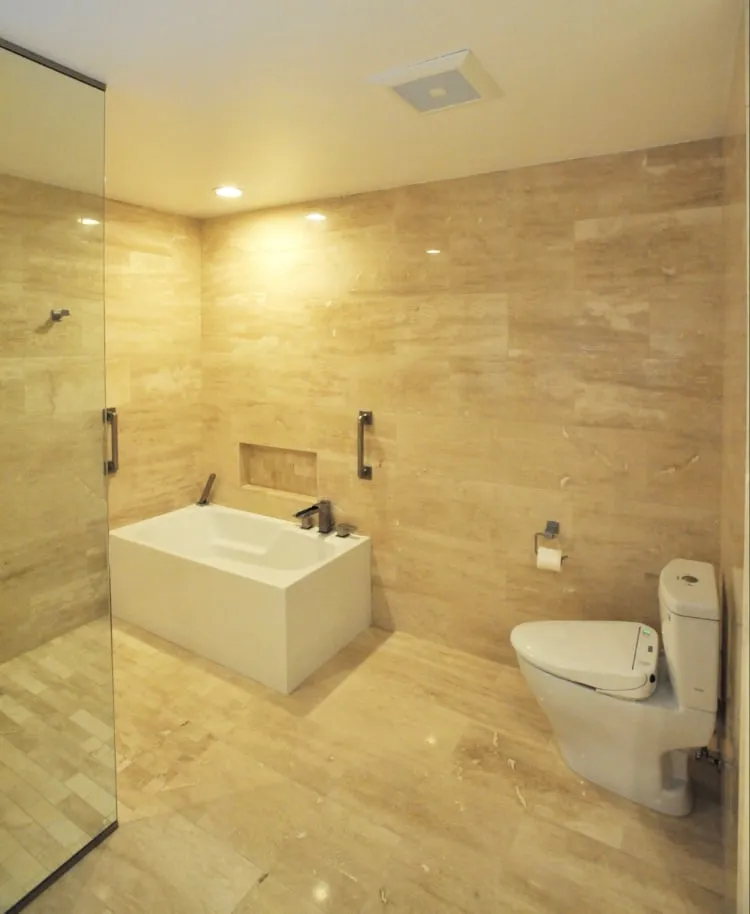 The finished bathroom: travertine walls and floor, a freestanding soaking tub in a tiled alcove with a recessed niche, and a frameless glass shower.