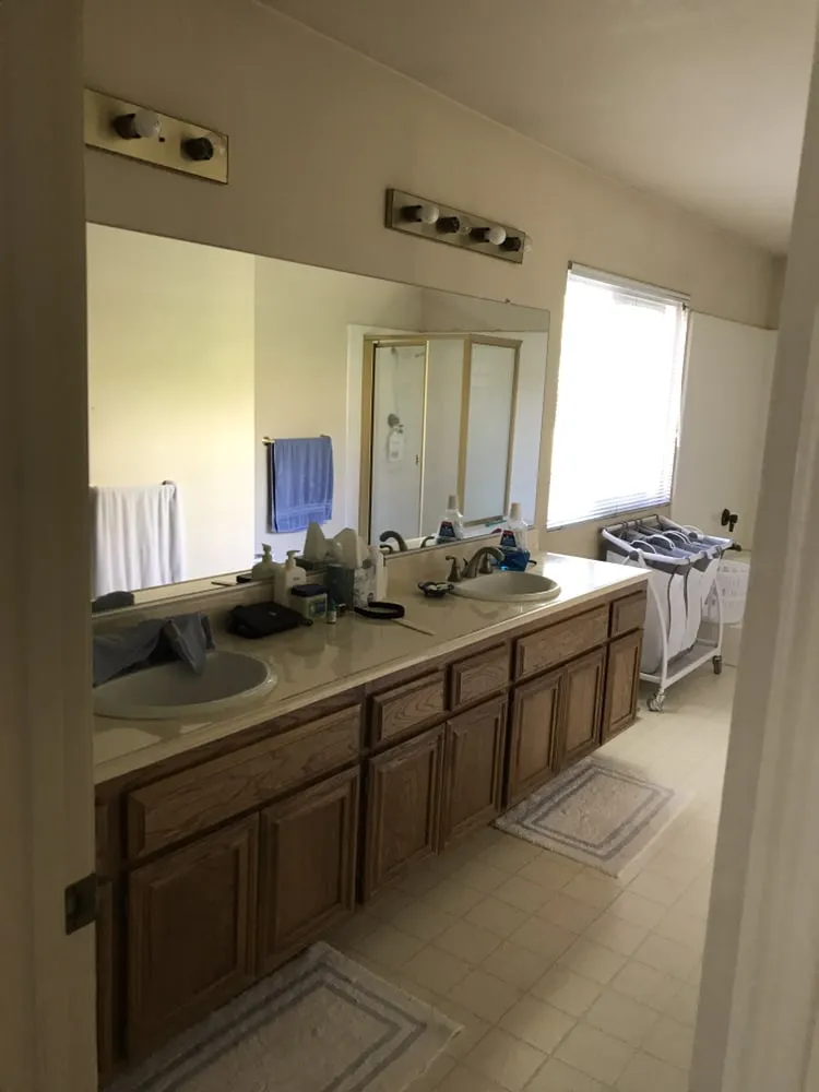 The bathroom before work: a long oak double vanity with a cultured-marble top, a full-width mirror, strip lighting and beige floor tile.