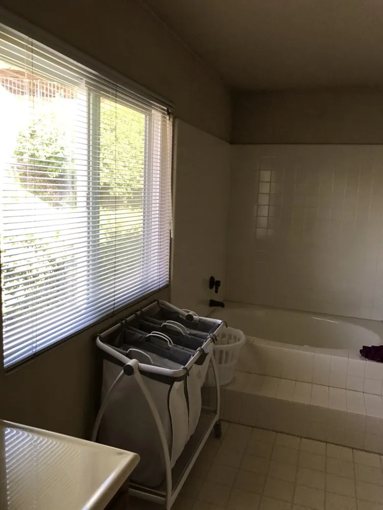 The same corner before work: a drop-in tub with a small-format tile surround beneath a blinded window, and dated floor tile.