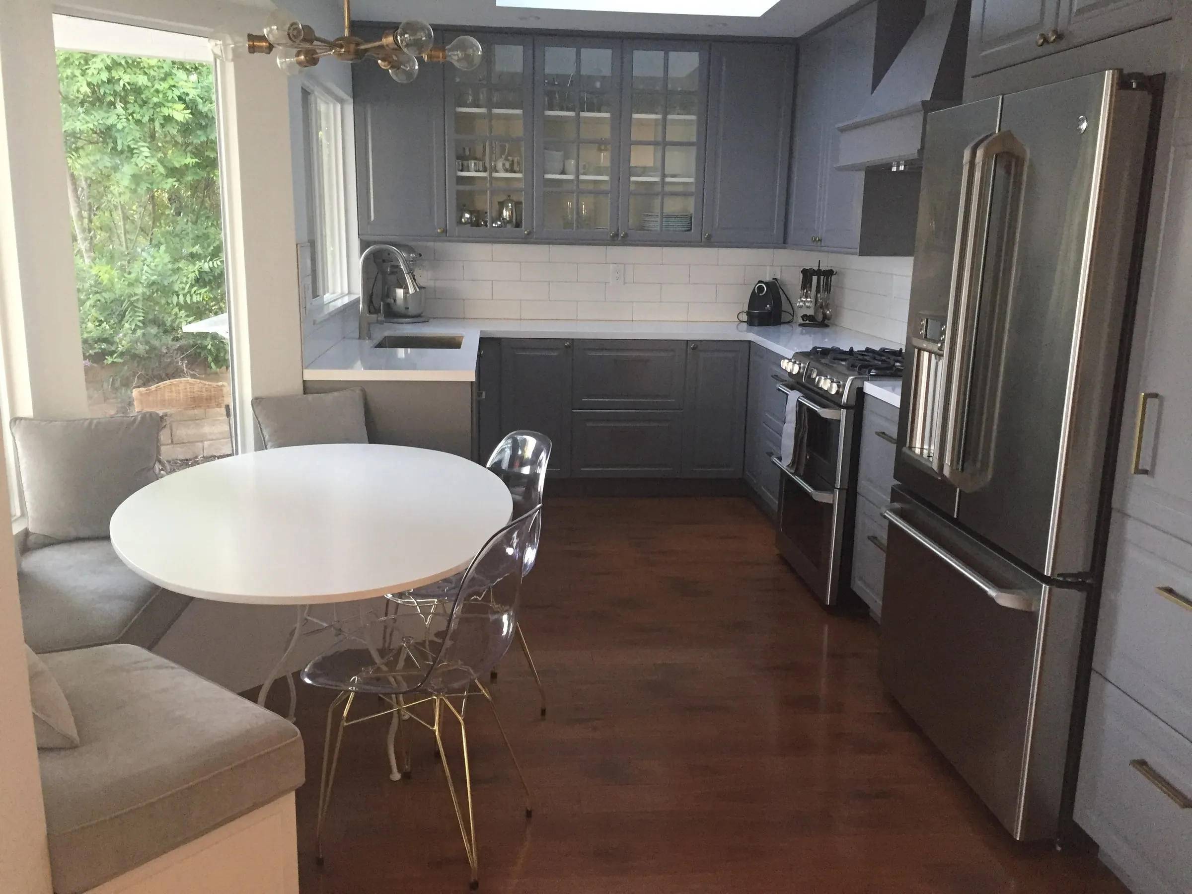 The kitchen after work: grey shaker cabinets with glass-front uppers, quartz counters, white subway tile, and a built-in banquette in the window bay.
